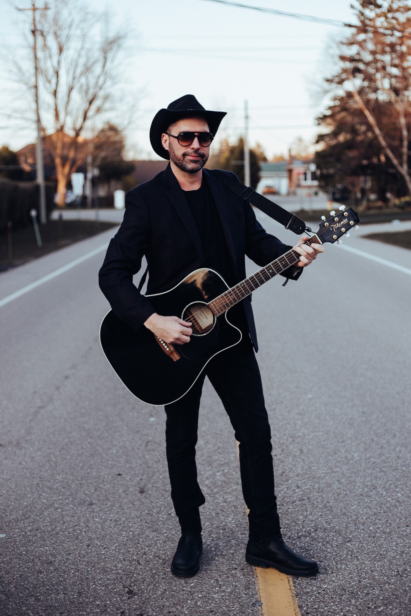 Lance Clyne in a cowboy hat and dark suit, holding his guitar outdoors