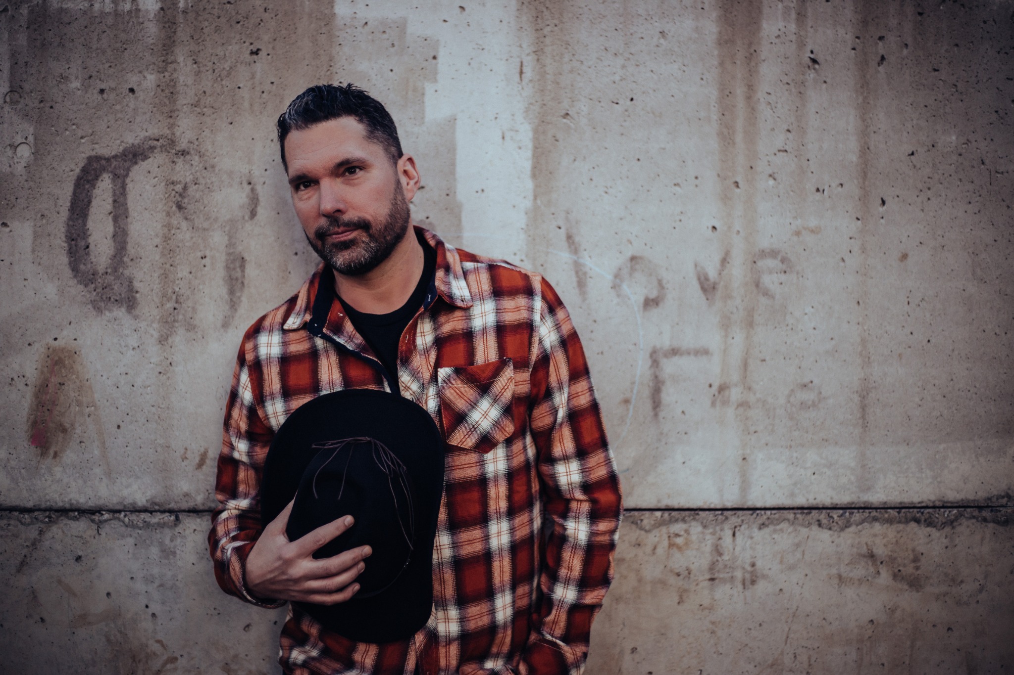 Promotional portrait in a plaid shirt beside a concrete wall